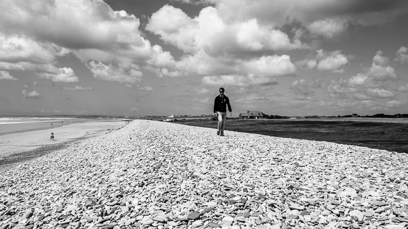 un homme marche sur un banc de galets, à droite la mer à gauche la terre