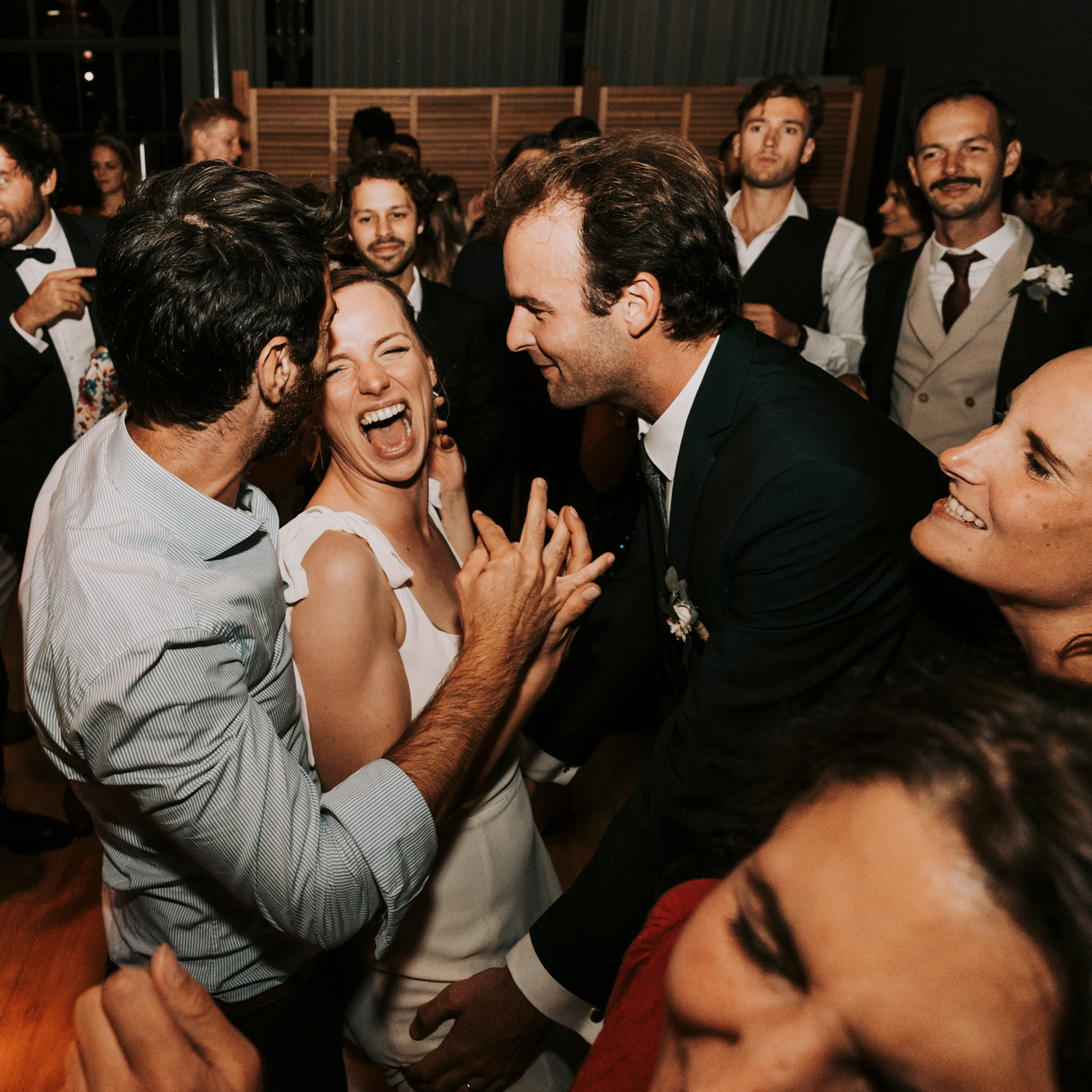 La mariée et le marié, entourés de leurs invités, rient et dansent joyeusement lors de la soirée de leur mariage à Rennes. La mariée, éclatante de bonheur dans sa robe blanche, partage un moment de complicité et de rires avec ses amis et son époux. Le marié, en costume sombre, se penche vers elle avec un sourire. Les invités les entourent, créant une atmosphère de fête et de célébration. La salle de réception est remplie d'énergie et de joie, capturant l'esprit festif de la soirée. Cette image illustre l'enthousiasme et l'allégresse qui marquent les célébrations de mariage.