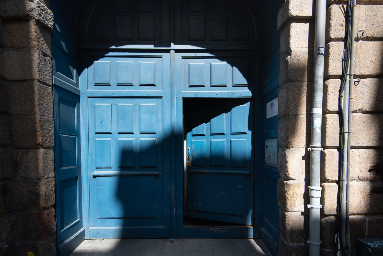 Vue de l'entrée du studio photo à Rennes avec ses grandes portes bleues emblématiques. La lumière du soleil crée des ombres artistiques sur les portes en bois, ajoutant du caractère à l'image. Cette photo illustre le charme et l'accueil chaleureux de l'espace où se déroulent mes séances photo