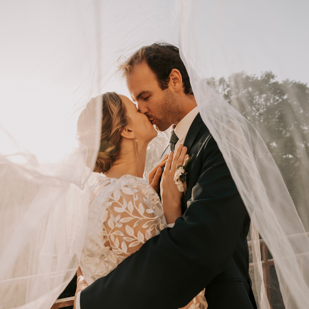 Un couple échangeant un tendre baiser sous le voile de la mariée lors d'une séance photo de mariage à Rennes. La mariée, dans une élégante robe en dentelle blanche avec des motifs floraux, et le marié, en costume sombre, se tiennent l'un contre l'autre, enveloppés par la douceur du voile. La lumière douce du soleil éclaire leur visage, créant une ambiance romantique et intemporelle. Cette image capture l'intimité et l'amour profond du couple, symbolisant les moments inoubliables d'un mariage.