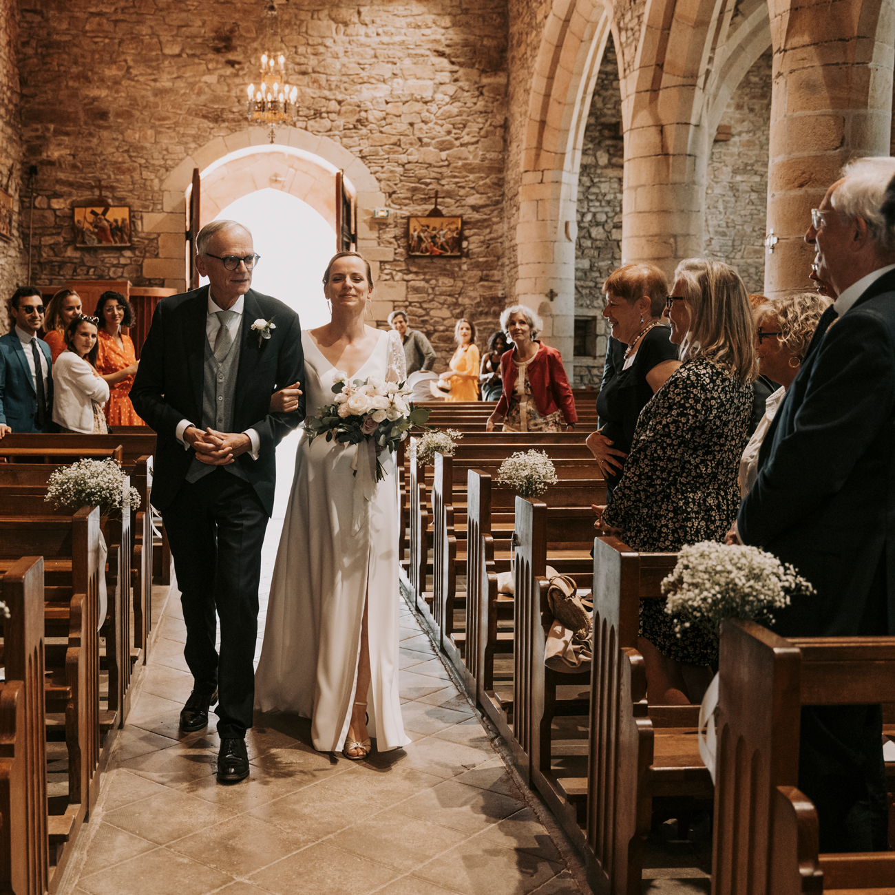 Une mariée et son père marchant dans l'allée centrale d'une église en pierre, entourés de leurs invités lors d'une cérémonie de mariage à Rennes. La mariée, dans une robe blanche élégante, tient un bouquet de fleurs blanches et vertes, tandis que son père, en costume sombre, l'accompagne avec un regard fier. Les invités, debout dans les bancs de l'église, regardent avec admiration et émotion. La lumière douce entrant par les portes ouvertes crée une ambiance chaleureuse et sacrée. Cette image capture le moment émouvant où la mariée fait son entrée, symbolisant le début de la cérémonie religieuse de mariage.