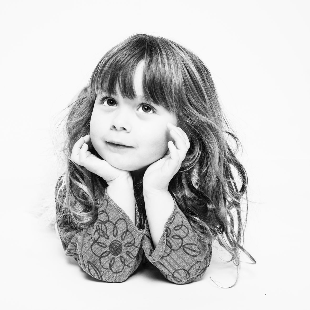 Portrait en noir et blanc d'une jeune fille lors d'une séance photo en studio à Rennes. La petite fille, avec ses cheveux bouclés et son visage encadré par ses mains, regarde vers le haut avec une expression rêveuse. Elle est vêtue d'un pull à motifs floraux, ce qui ajoute une touche douce et enfantine à la photo. L'arrière-plan blanc pur met en valeur les détails et les nuances de son visage, capturant son innocence et sa curiosité. Cette image illustre parfaitement l'art de la photographie de portraits d'enfants en studio.