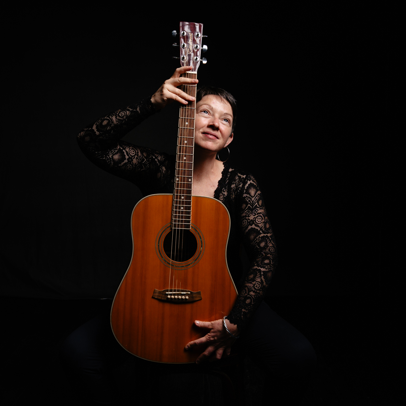 Portrait en studio d'une femme tenant une guitare acoustique lors d'une séance photo d'estime de soi à Rennes. Vêtue d'un haut en dentelle noire, elle pose avec confiance et charme, la guitare positionnée devant elle. La lumière douce éclaire son visage tandis qu'elle regarde vers le haut, créant une ambiance chaleureuse et introspective. Le fond noir met en valeur son expression joyeuse et l'instrument, capturant l'essence de sa passion musicale.