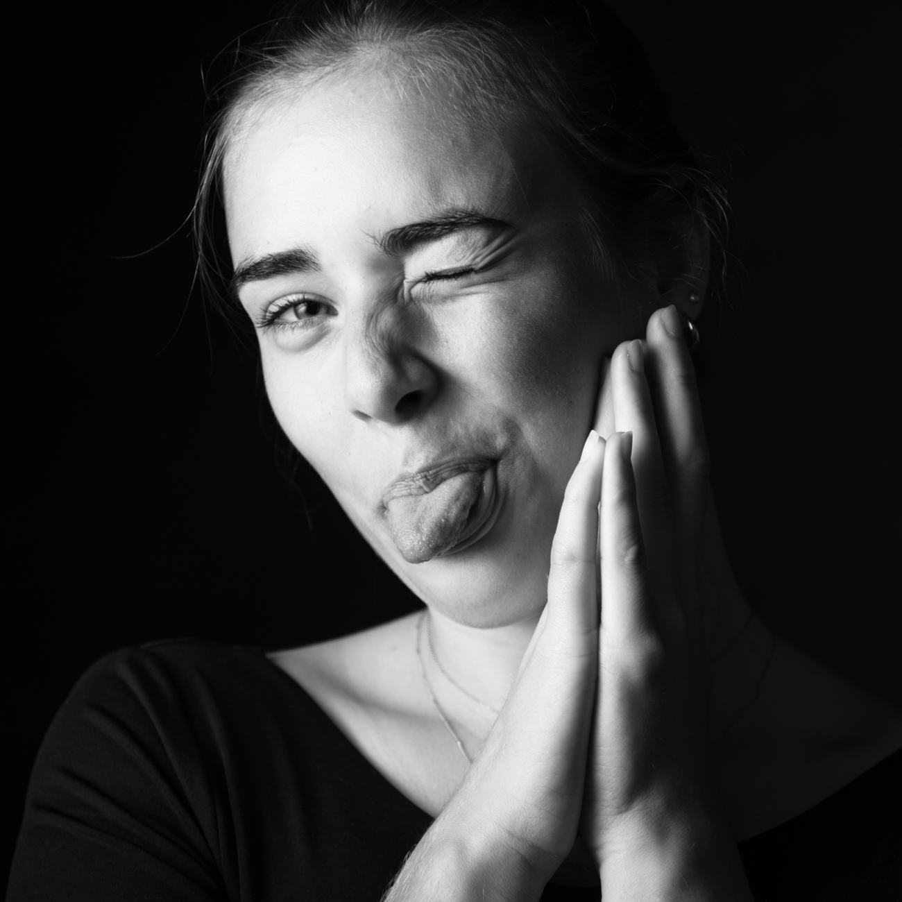 Portrait en noir et blanc d'une jeune femme tirant la langue et clignant des yeux lors d'une séance photo d'estime de soi à Rennes en studio. La femme affiche une expression joyeuse et joueuse, posant ses mains sur ses joues. La lumière met en valeur les traits de son visage contre un fond sombre, capturant un moment de spontanéité et de confiance. Cette image illustre parfaitement l'authenticité et l'énergie positive que l'on peut ressentir lors d'une séance photo pour booster l'estime de soi.