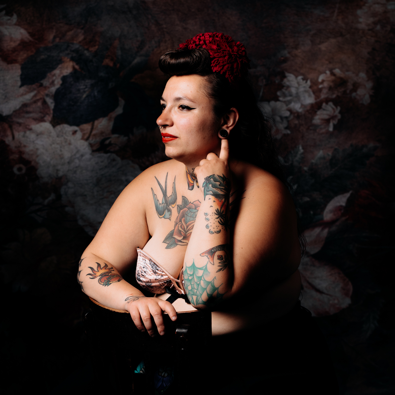 Portrait d'une femme tatouée lors d'une séance photo en studio à Rennes. Elle est assise avec une posture élégante, portant un soutien-gorge fleuri et un accessoire rouge dans les cheveux. Son regard est tourné vers le côté, exprimant à la fois force et sérénité. La lumière met en valeur ses tatouages détaillés, notamment des motifs floraux et marins, contrastant avec le fond sombre et texturé. Cette image illustre parfaitement l'expression de la confiance en soi et de l'authenticité personnelle lors d'une séance photo d'estime de soi.