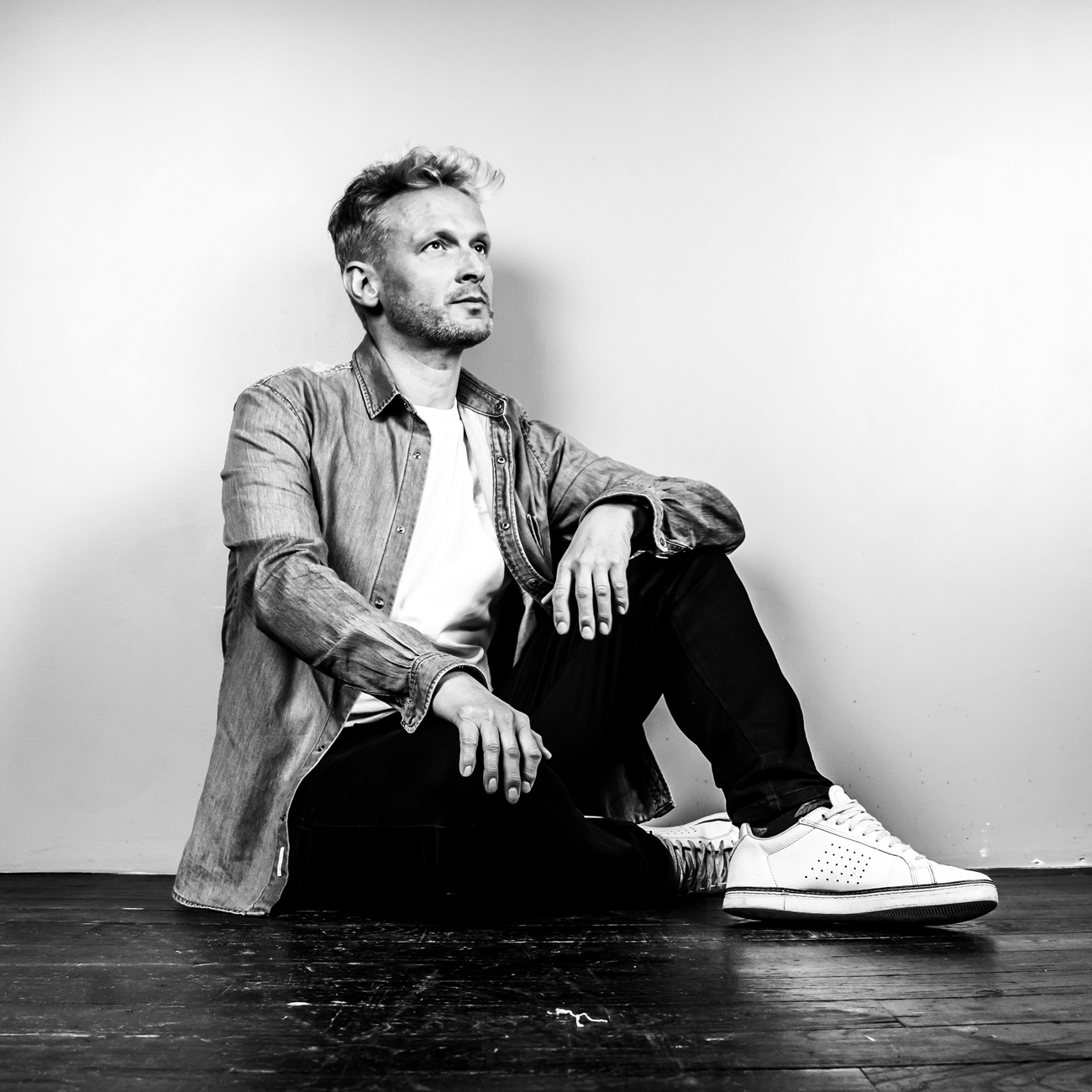 Portrait en noir et blanc d'un homme assis sur le sol lors d'une séance photo en studio à Rennes. Vêtu d'une chemise en jean, d'un t-shirt blanc et de jeans noirs, il pose de manière décontractée avec un regard pensif tourné vers la lumière. Ses baskets blanches contrastent avec le sol sombre, ajoutant une touche moderne et dynamique à l'image. L'éclairage met en valeur les détails de son expression et sa posture, capturant une atmosphère de réflexion et de sérénité. Cette photo illustre l'art de la photographie de portrait en studio, mettant en avant la personnalité et la confiance du sujet