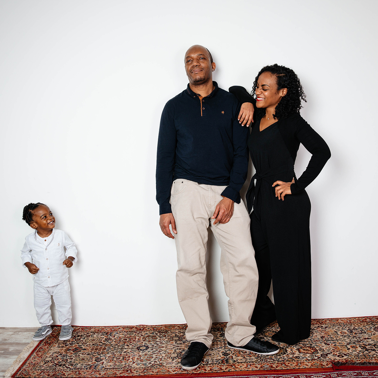 Une famille posant lors d'une séance photo en studio à Rennes. Le père, la mère, et leur jeune enfant se tiennent debout sur un tapis, chacun affichant des sourires rayonnants. Le père, vêtu d'une chemise bleue et d'un pantalon beige, regarde droit devant, tandis que la mère, en tenue noire élégante, sourit affectueusement à leur enfant vêtu de blanc, qui les regarde avec joie. La scène capture l'affection et la complicité familiale dans un cadre simple et lumineux. Cette photo illustre parfaitement les liens familiaux et la chaleur d'une séance photo de famille.