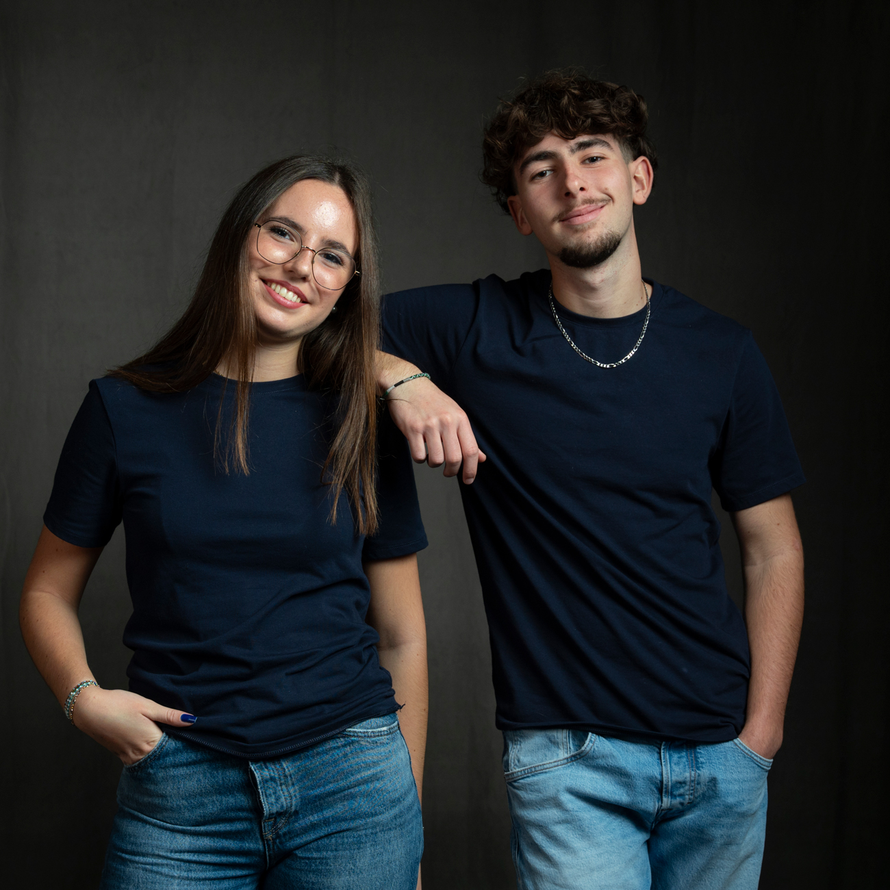 Frère et sœur posant ensemble lors d'une séance photo en studio à Rennes. Tous deux habillés en jeans et t-shirts bleu foncé assortis, ils sourient et montrent une complicité naturelle. Le frère, adossé légèrement contre sa sœur, affiche un sourire confiant, tandis que la sœur, souriante, pose avec aisance les mains dans les poches. La simplicité de leurs tenues et l'arrière-plan sombre mettent en valeur leurs expressions et leur lien familial. Cette photo illustre parfaitement la capture des portraits familiaux dans un cadre studio professionnel.