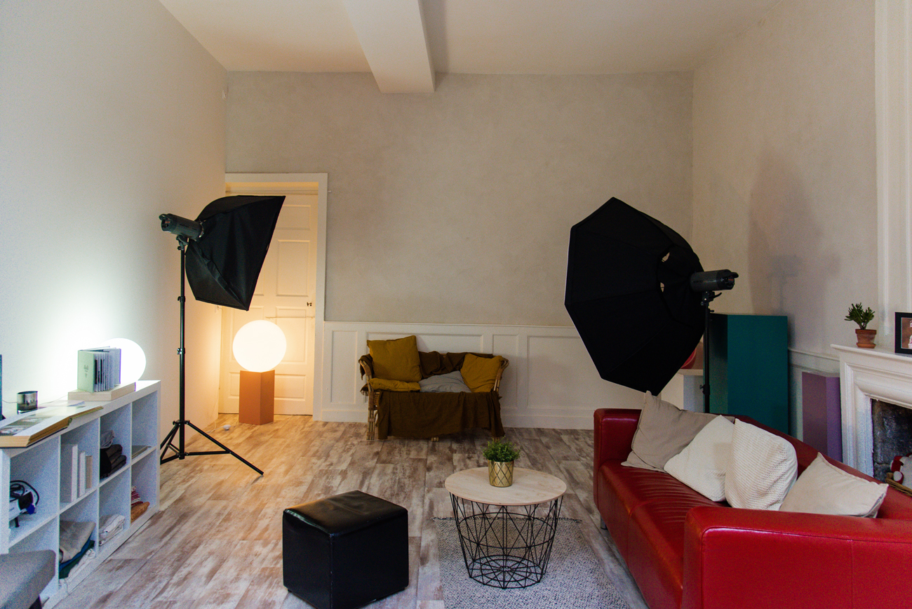 Vue intérieure du studio photo à Rennes prêt pour une séance photo professionnelle. Le studio est équipé de lumières de studio modernes et d'un canapé rouge confortable, créant une atmosphère accueillante et chaleureuse. Des accessoires et des meubles élégants sont disposés autour de l'espace, offrant des options variées pour les séances photo familiales et individuelles. Cette photo montre le cadre professionnel et convivial où les séances se déroulent, mettant en valeur l'attention portée aux détails et au confort des clients